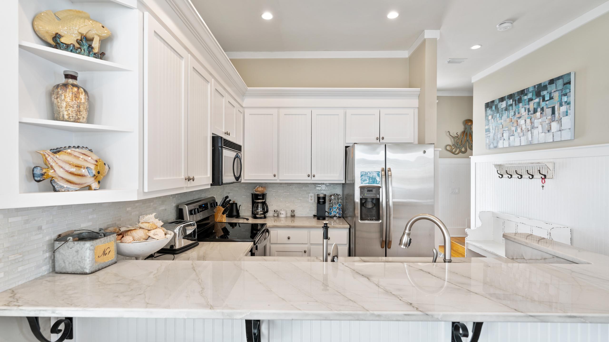 A bright kitchen with white shaker cabinets, marble countertops, and stainless steel appliances, including a large refrigerator. Decorative items like shells and a fish sculpture adorn open shelving.