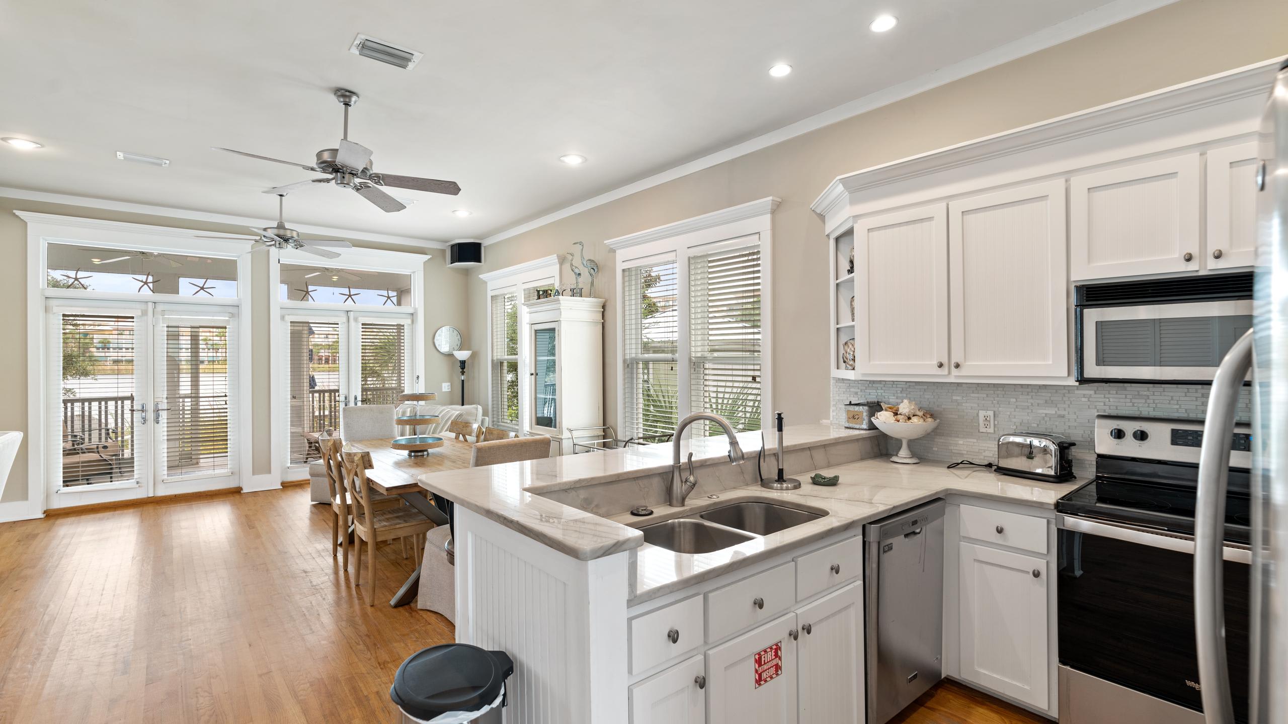 An open-concept kitchen with white cabinets and marble countertops, overlooking a dining area with a wooden table and chairs. Large glass doors lead to an outdoor space, and ceiling fans are visible.