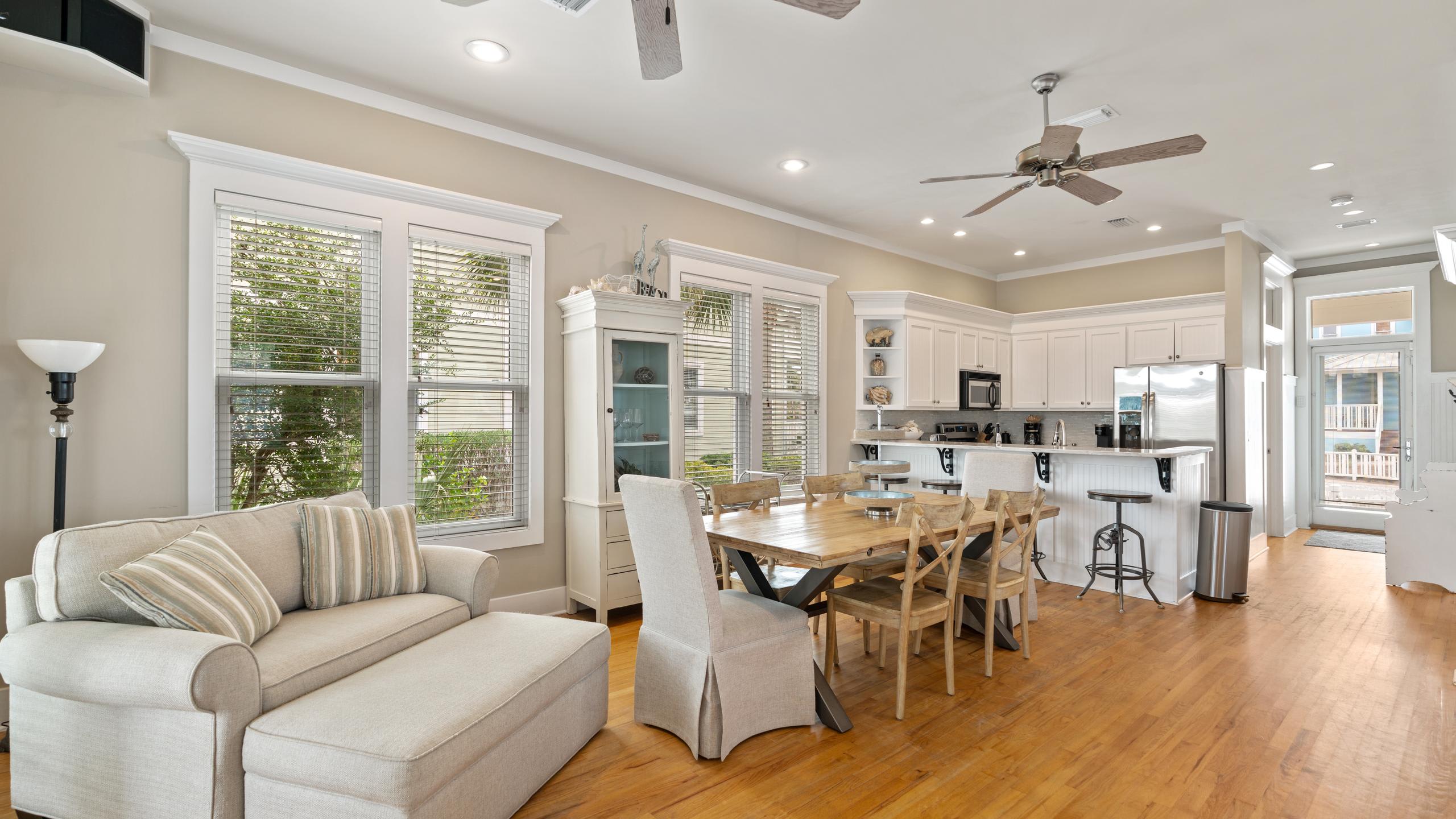 An open-concept dining area with light hardwood floors and beige walls includes a rustic wooden dining table, a chaise lounge, and views into a white kitchen with a breakfast bar and stainless steel appliances.