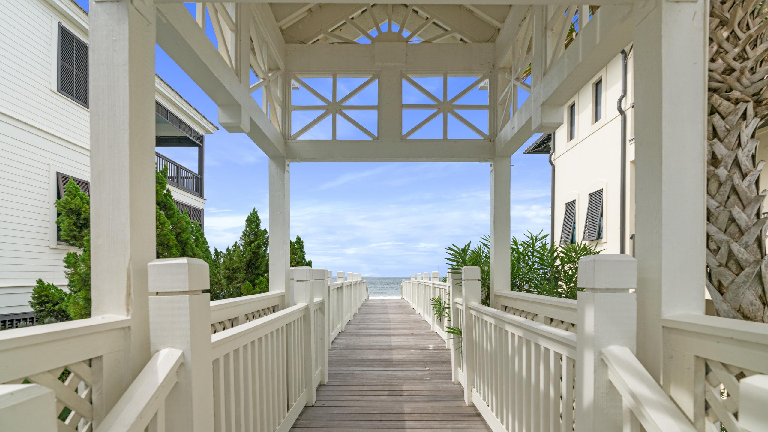 A picturesque wooden boardwalk leads through a white archway towards a distant view of the ocean under a clear blue sky, flanked by lush green foliage and white multi-story buildings.