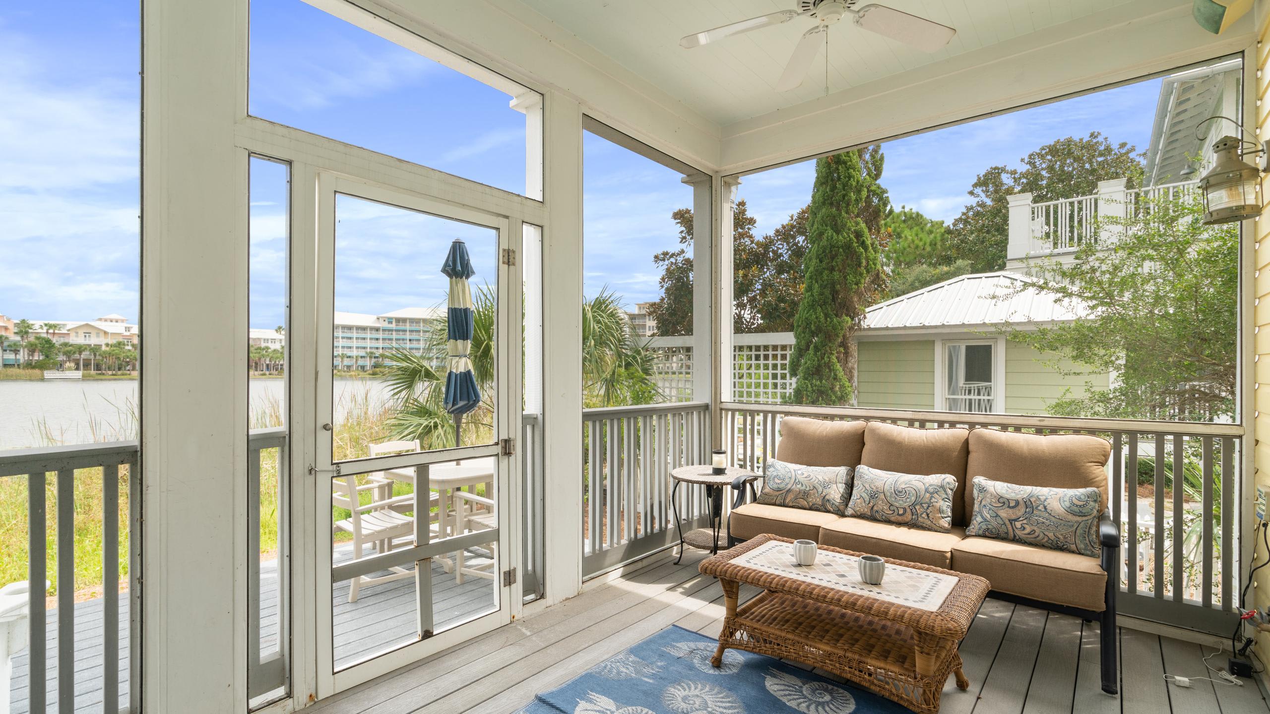 A serene screened-in porch features a comfortable brown sofa with patterned pillows, a wicker coffee table, and a blue rug, offering a picturesque view of a lake and distant buildings under a clear sky.