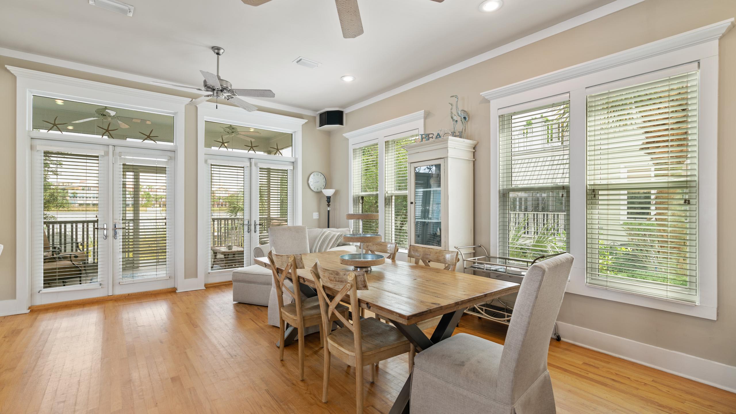 A spacious dining area features a rustic wooden table with seating for six, light hardwood floors, and large windows with blinds, leading to a set of French doors that open to an outdoor space.
