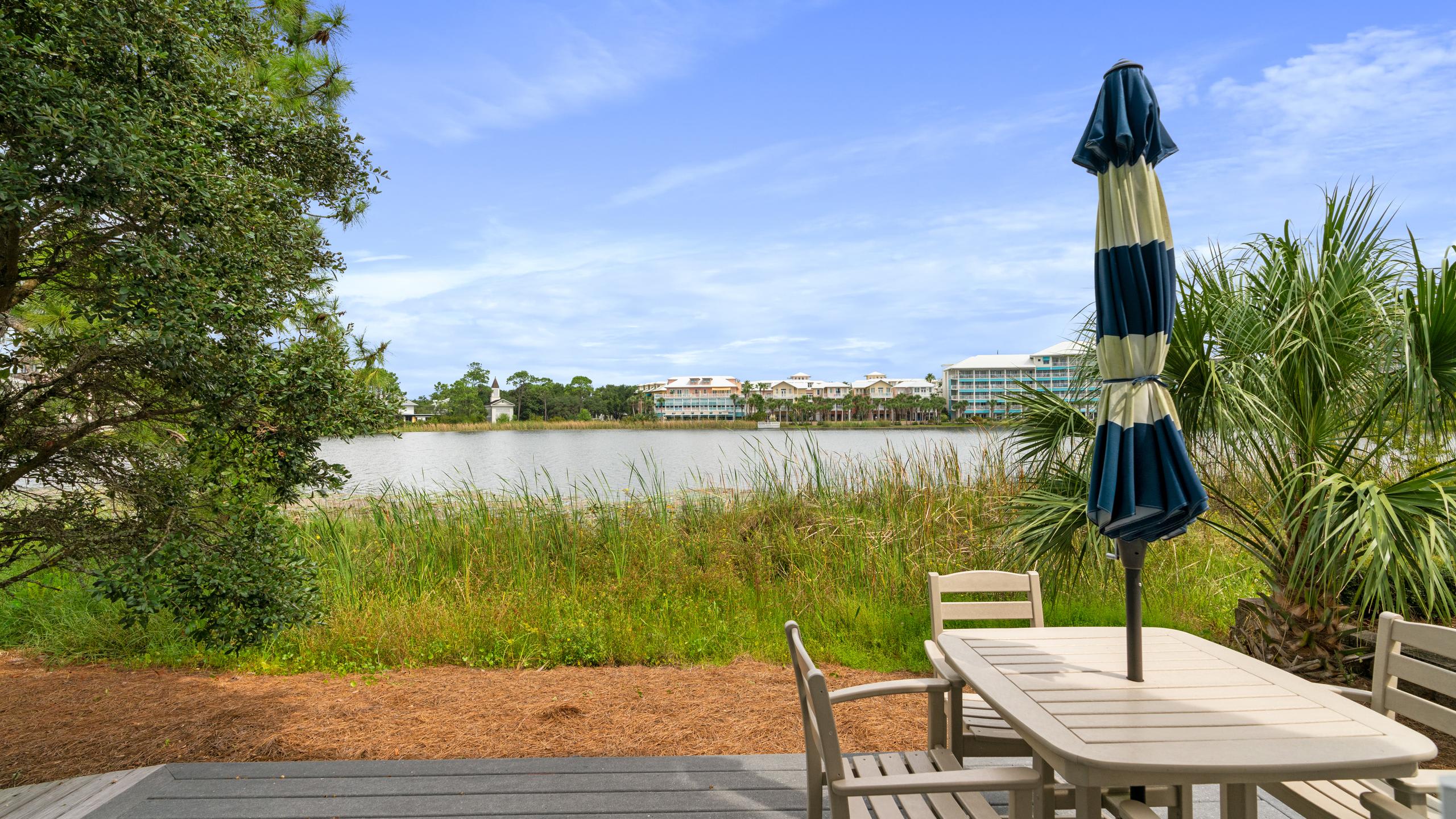 An outdoor deck with a white patio table and chairs, shaded by a blue and white striped umbrella. The deck overlooks a serene lake with lush green reeds and distant buildings under a blue sky.