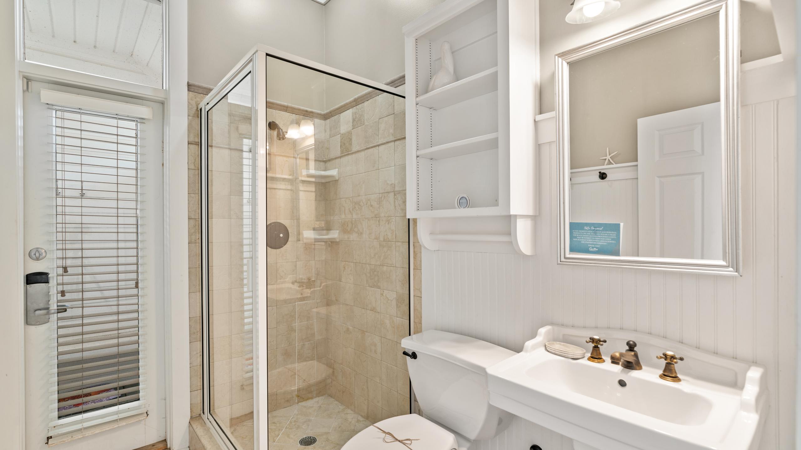 A compact bathroom with white beadboard walls, a pedestal sink with brass fixtures, and a glass-enclosed shower with beige tiled walls. A medicine cabinet and small shelf are mounted above the toilet.
