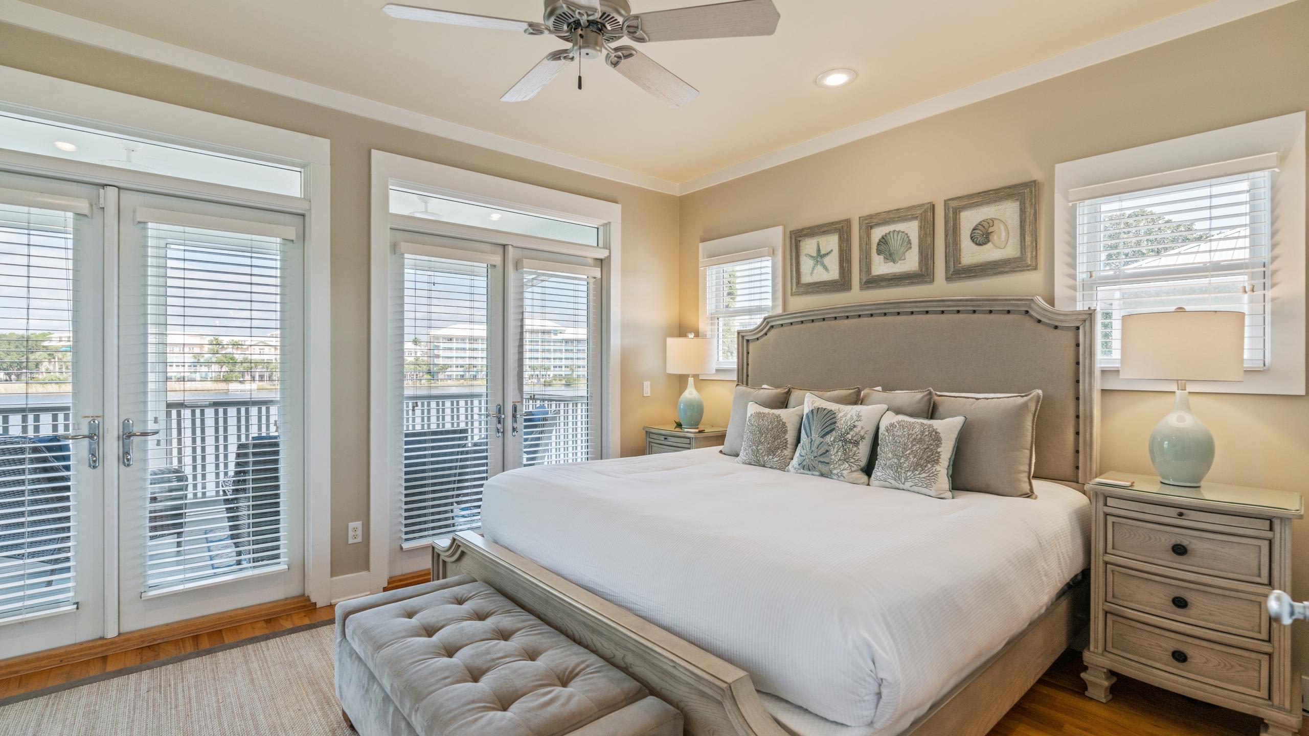 Elegant bedroom with a large king-sized bed, two nightstands with lamps, and multiple windows with white blinds. French doors lead to a balcony overlooking a lake.