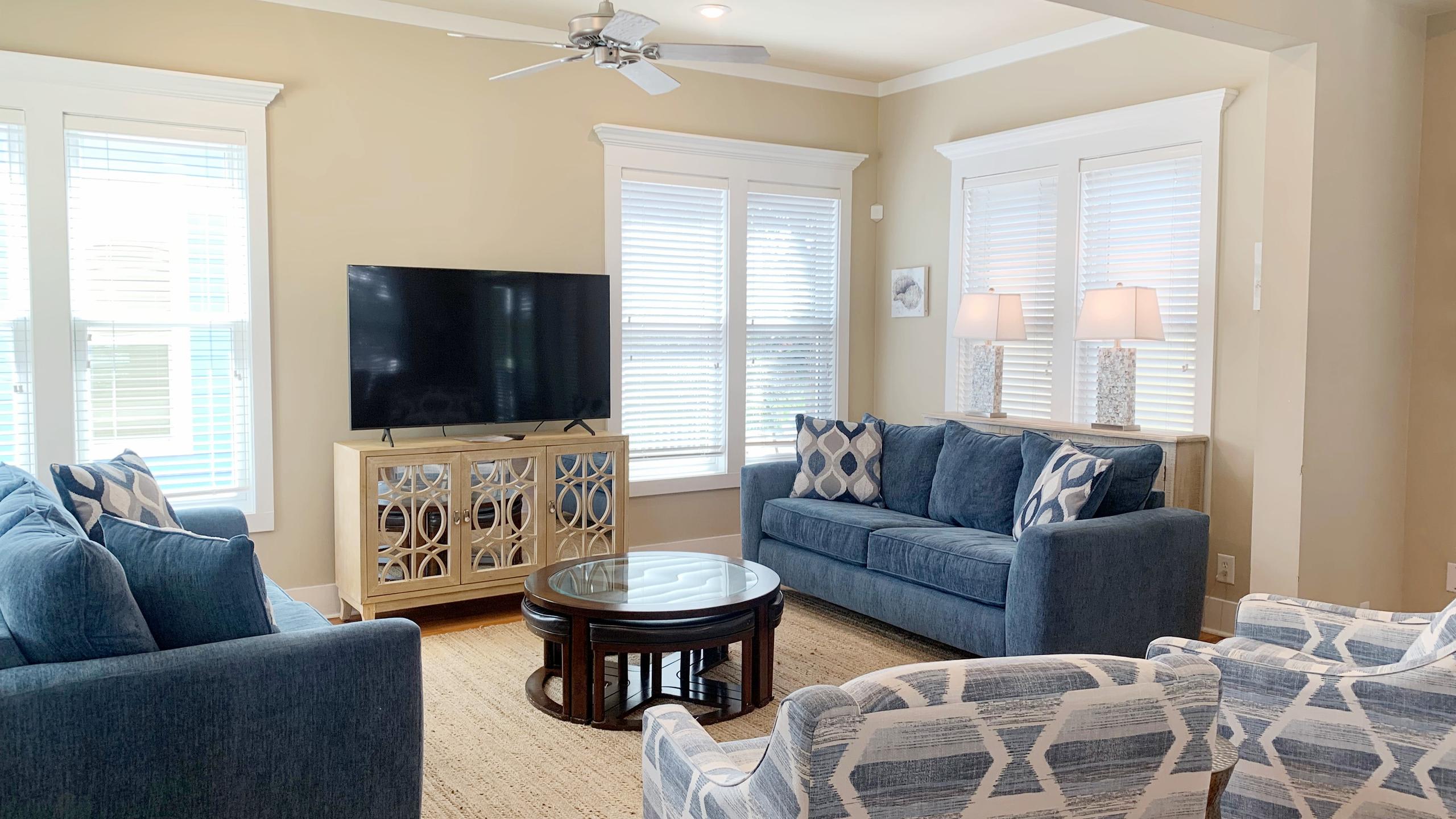 A bright living room features two blue sofas with patterned pillows, a large wall-mounted TV on a decorative console, and a round dark wood coffee table, all set on a textured natural rug.