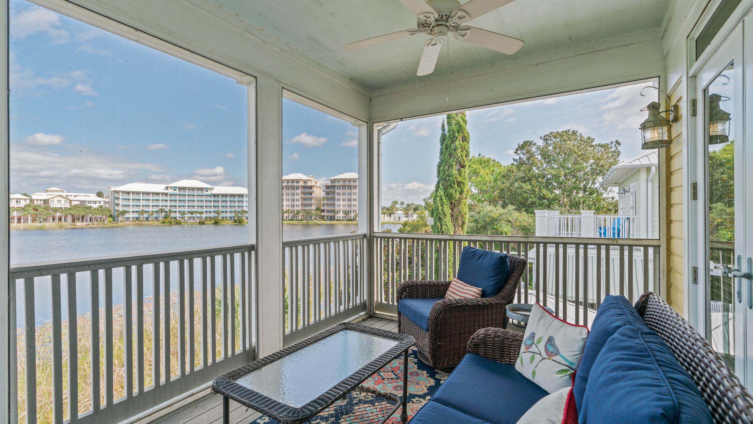 Screened porch with dark wicker furniture, blue cushions, and a glass-top coffee table, offering a wide view of a lake and distant buildings. A ceiling fan hangs overhead.
