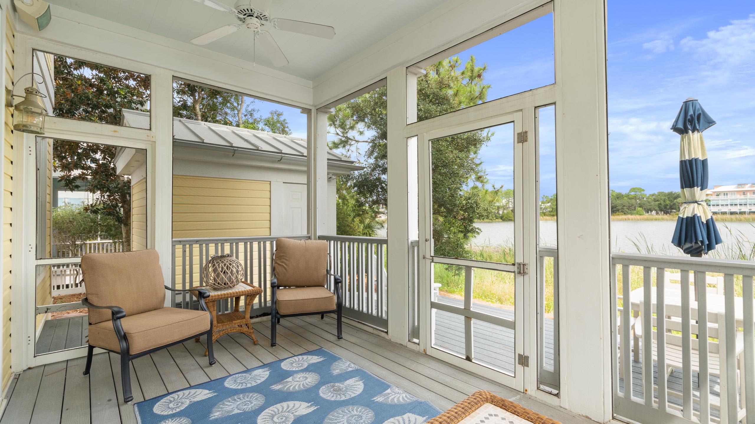 A screened-in porch with two comfortable armchairs and a small side table, featuring a blue rug with a shell pattern. The porch offers views of the surrounding greenery and a lake in the distance.