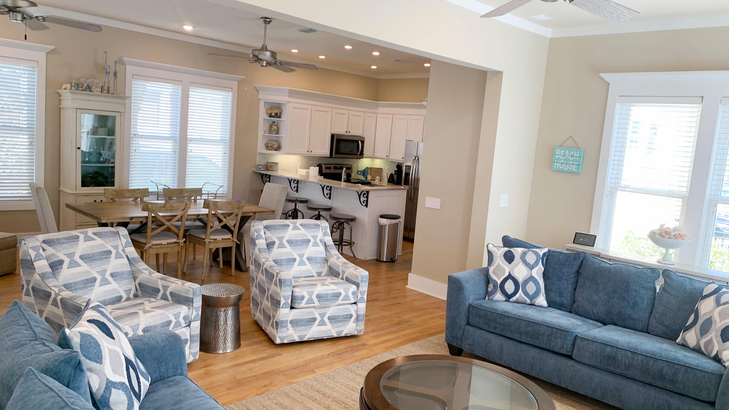 An open-concept living area showcases two blue sofas and two patterned armchairs around a coffee table, with a view into a bright kitchen featuring white cabinets, stainless steel appliances, and a breakfast bar.