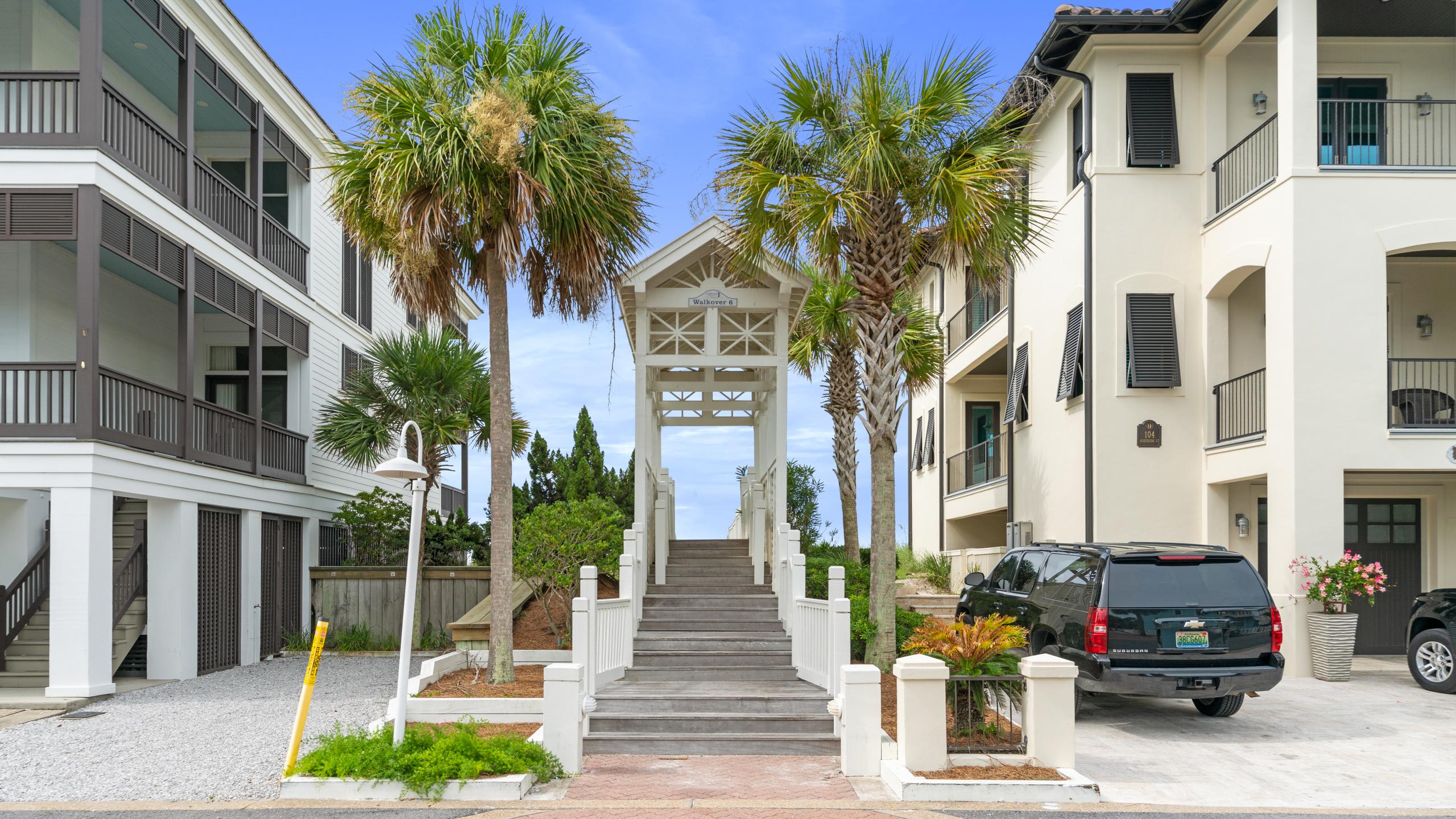 A grand wooden staircase with white railings ascends to a covered archway, leading to the beach, flanked by tall palm trees and multi-story vacation homes with dark shutters.