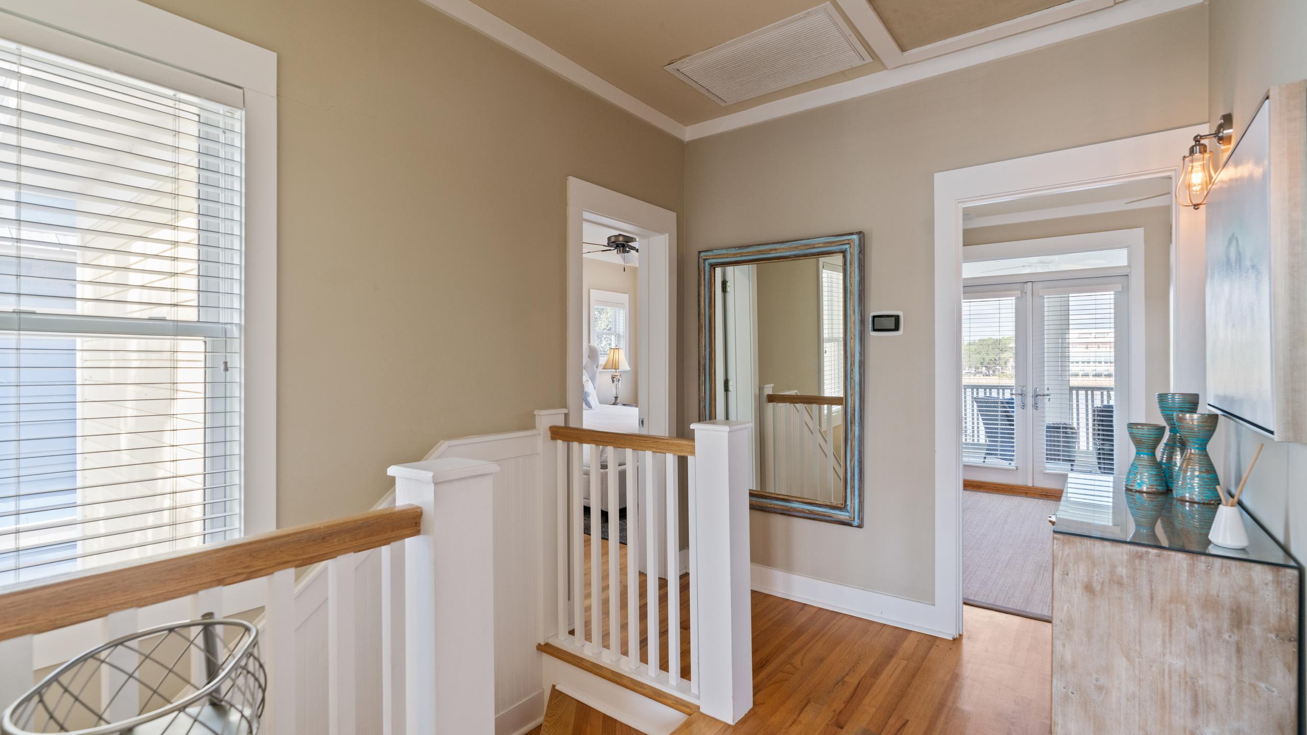 A bright upper-level landing features light hardwood floors, white railings, and a large distressed blue mirror, with glimpses into other rooms through open doorways and natural light from a window.