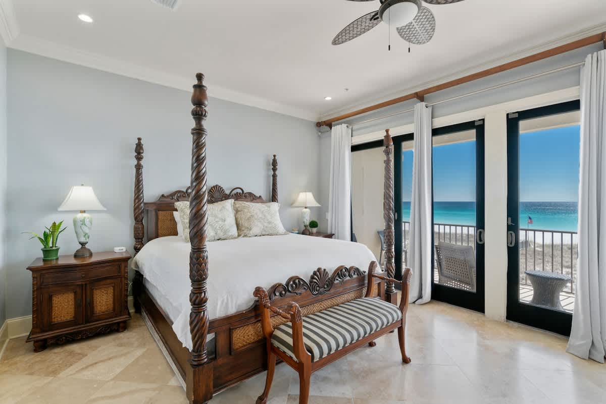 A spacious bedroom with light blue walls and travertine flooring, featuring a dark wood four-poster bed with white bedding, and large glass doors leading to a balcony with panoramic ocean views.