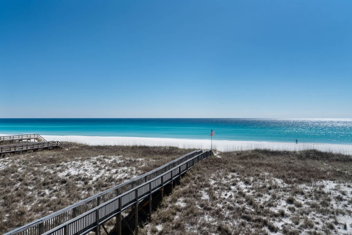 Expansive view of a pristine white sand beach with turquoise ocean waters under a clear blue sky. A long wooden boardwalk stretches across the coastal dunes towards the water, with an American flag visible near the beach.