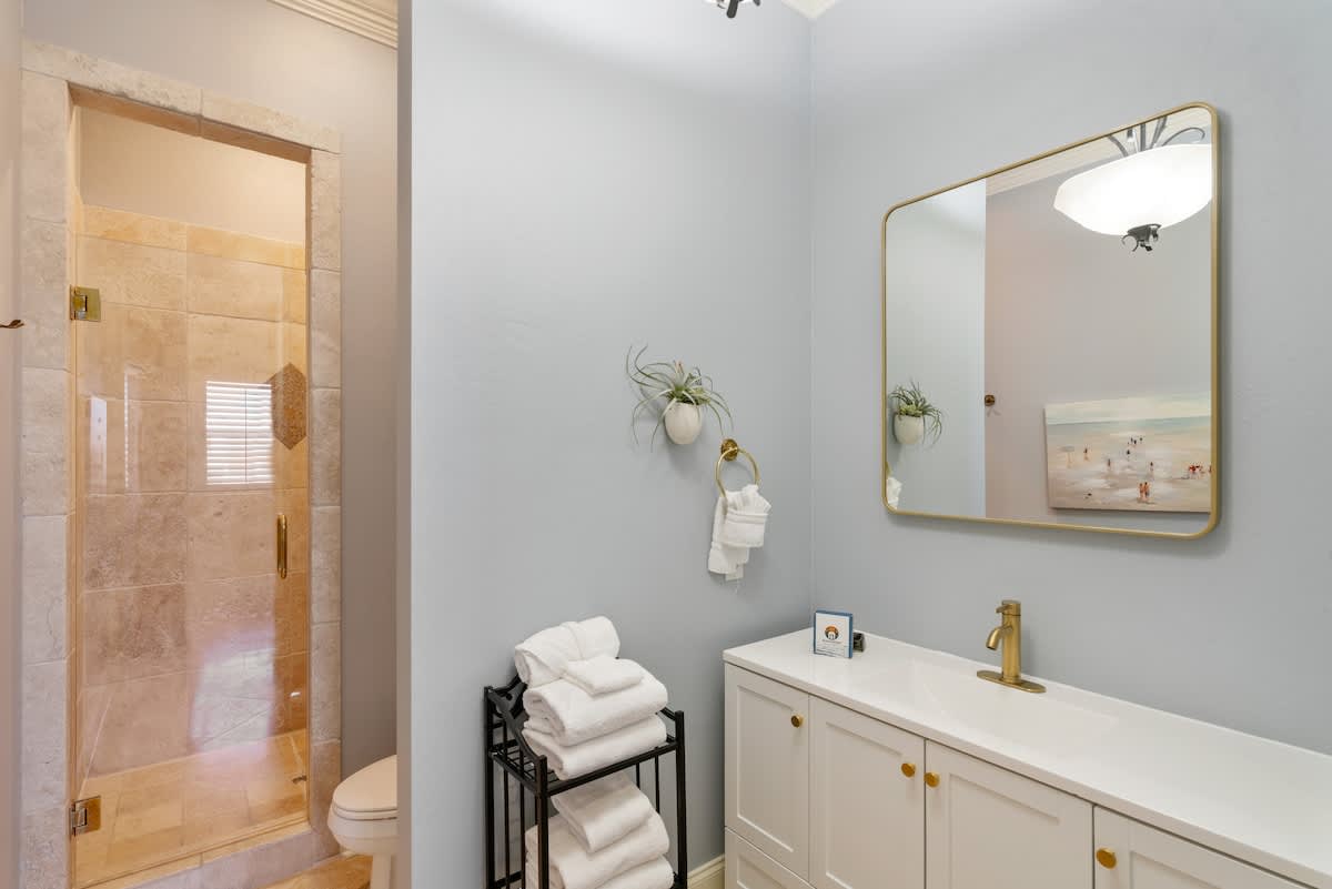 Light blue bathroom featuring a white single vanity with gold fixtures and a large rectangular mirror. A tiled walk-in shower with a glass door is visible, alongside a black metal shelf holding neatly folded white towels.