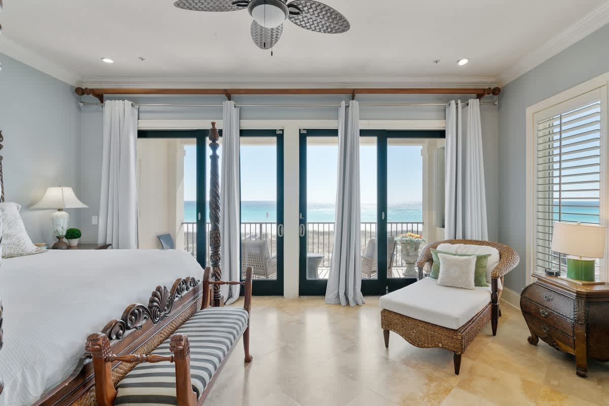 A luxurious bedroom with light blue walls and travertine flooring, featuring a dark wood four-poster bed, a ceiling fan, and floor-to-ceiling glass doors opening to a balcony with ocean views.