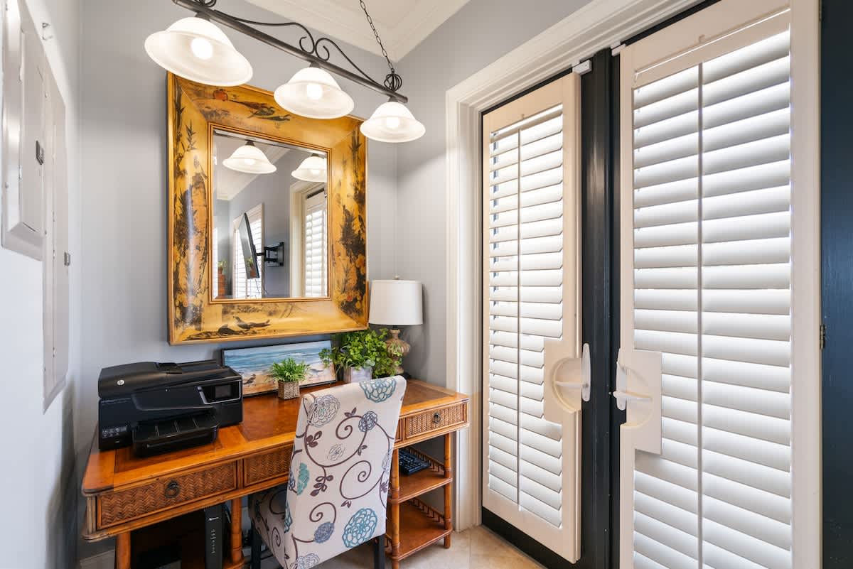 A dedicated workspace within a bedroom, featuring a wooden desk with a printer, a patterned chair, and a large gold-framed mirror reflecting the room. Double doors with plantation shutters lead to an exterior space.