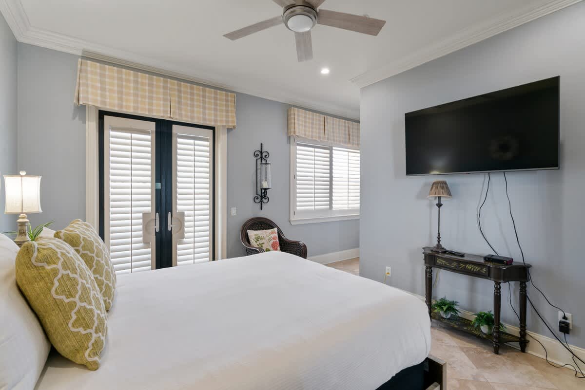 View of a bedroom with light blue walls and travertine tile flooring, showcasing a queen-sized bed with white bedding and yellow accent pillows. A wall-mounted TV, a decorative console table, and a wicker chair are also present.