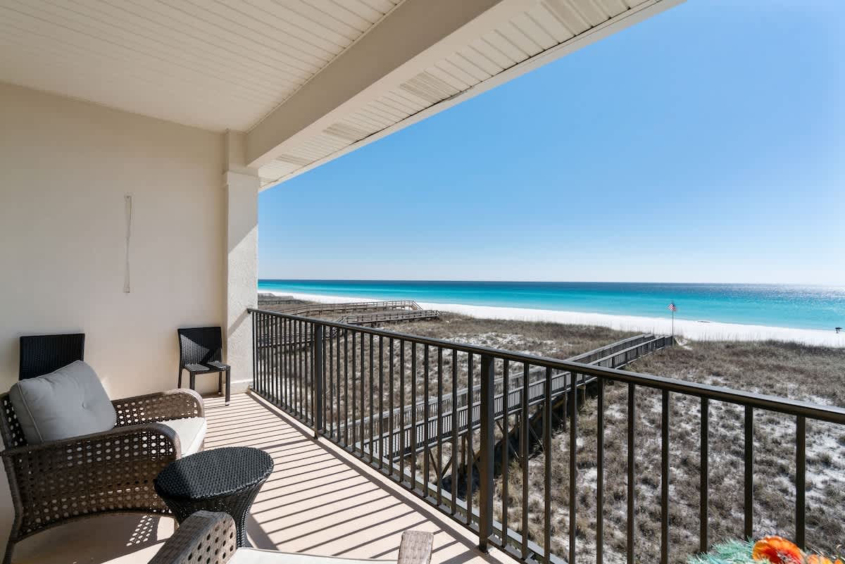Spacious covered balcony with black metal railing, offering panoramic views of the white sand beach, turquoise ocean, and a wooden boardwalk winding through coastal dunes. Wicker chairs and a small table furnish the outdoor space.