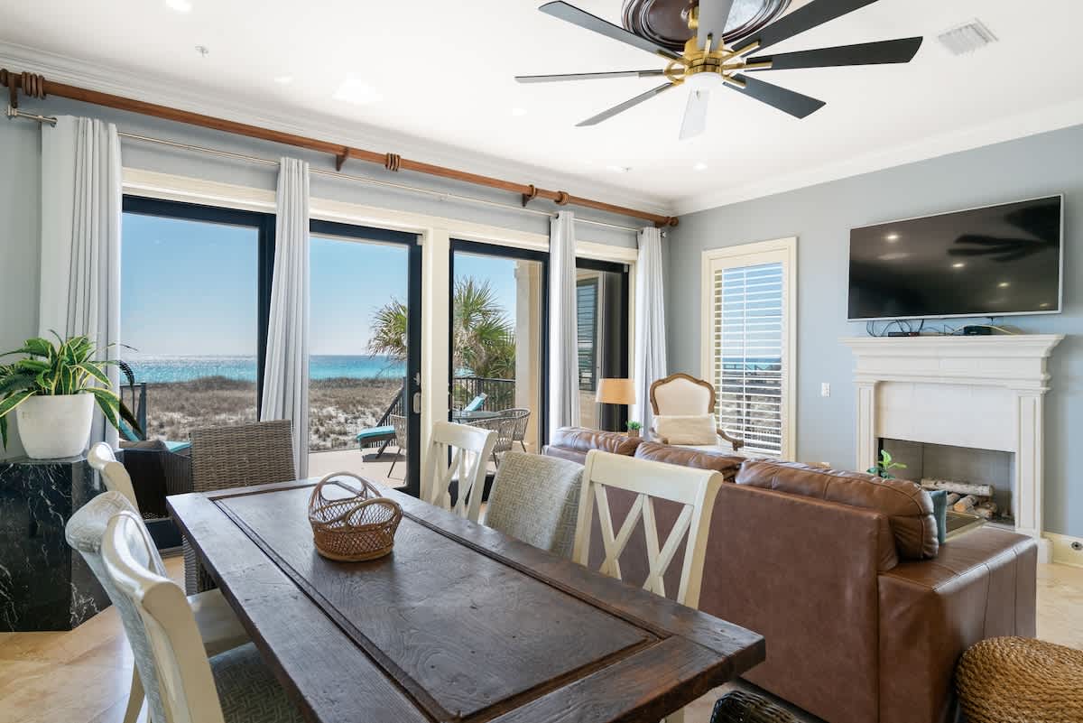 A bright open-concept living and dining area with a dark wood dining table, brown leather sectional, and large sliding glass doors offering panoramic beach and ocean views. A ceiling fan hangs above.