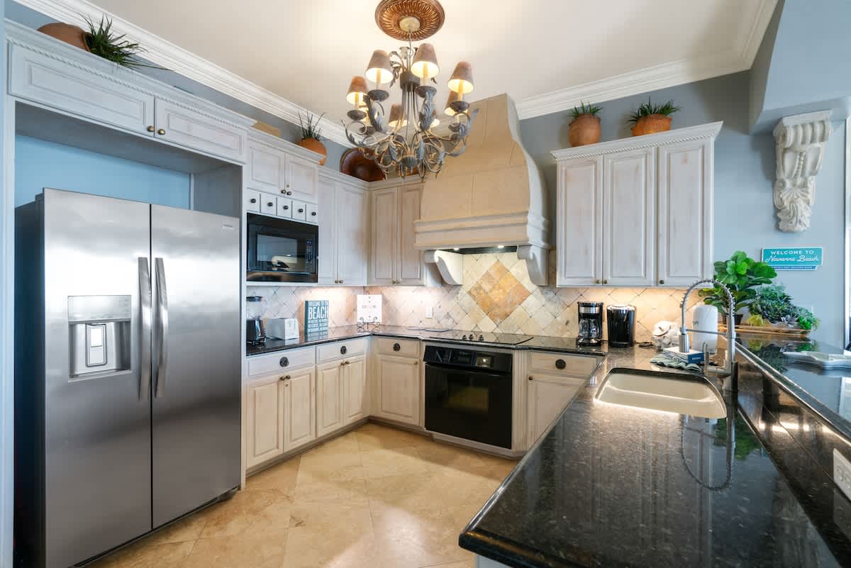 A detailed view of the kitchen showing light-colored cabinets, dark granite countertops, a tiled backsplash, stainless steel refrigerator, microwave, and oven, with a decorative range hood.