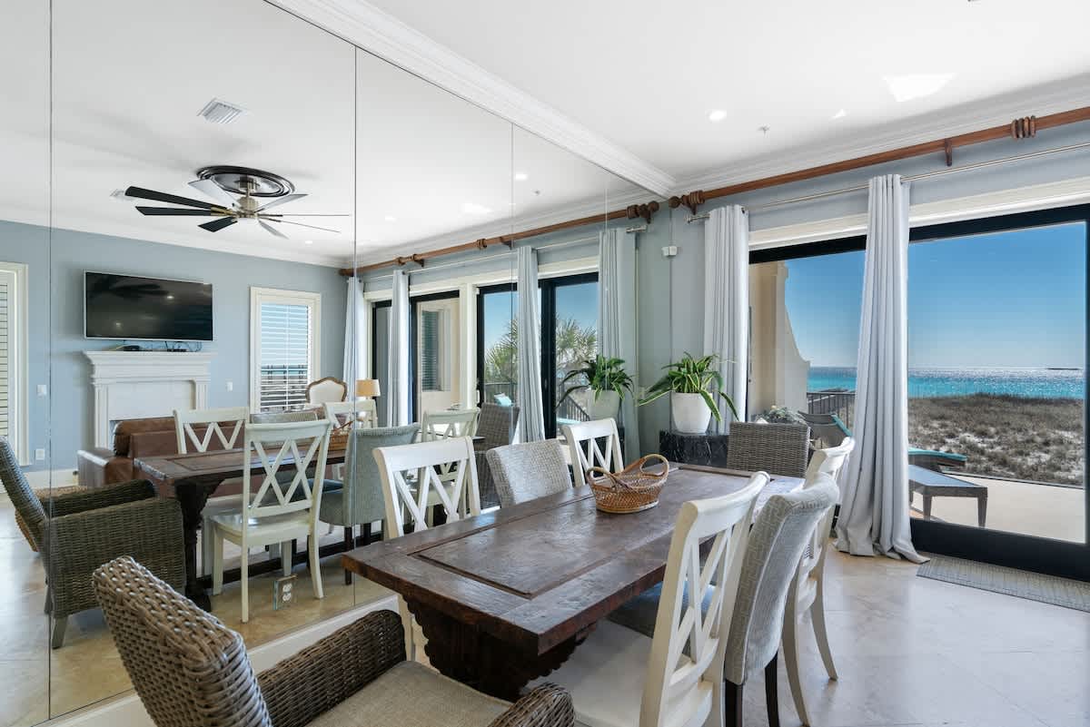 A bright dining area with a large wooden table and mixed seating, featuring a mirrored wall and floor-to-ceiling windows that provide stunning views of the beach and ocean.