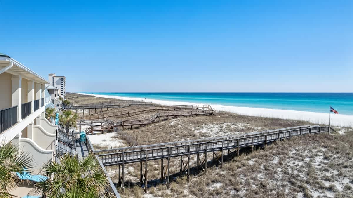 An elevated view from a balcony overlooking a vast expanse of white sand beach, turquoise ocean, and a series of wooden boardwalks winding through the dunes, with an American flag visible.
