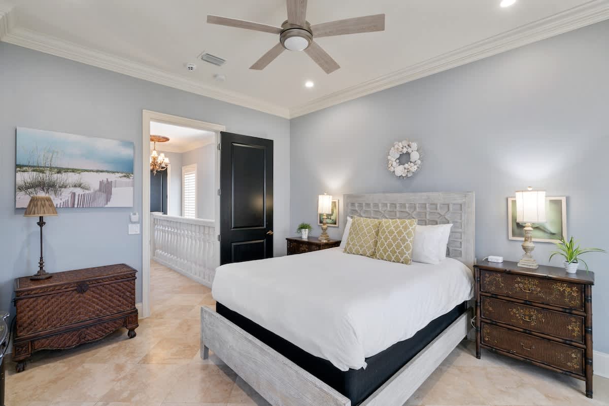 Serene bedroom featuring a queen-sized bed with a light wood headboard and white bedding, flanked by dark wood nightstands with lamps. A large beach-themed painting adorns the light blue wall, complemented by a ceiling fan and tile flooring.