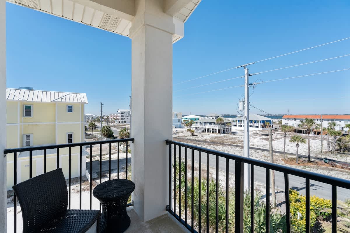 Small private balcony with black metal railing, offering a view of the street, neighboring houses, and palm trees. A black wicker chair and a small black table provide a cozy seating area.