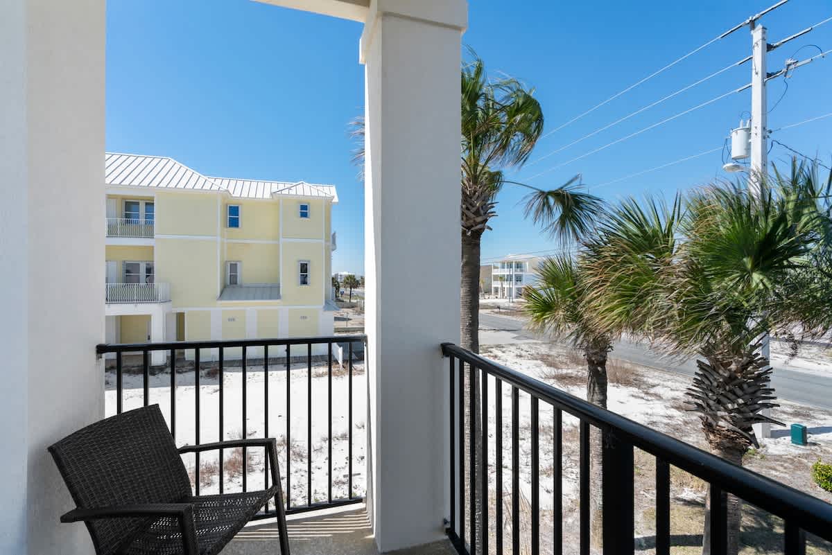 A sunny outdoor balcony with a black metal railing and a dark woven chair, offering a view of a sandy area, lush palm trees, and a light yellow building under a clear blue sky.