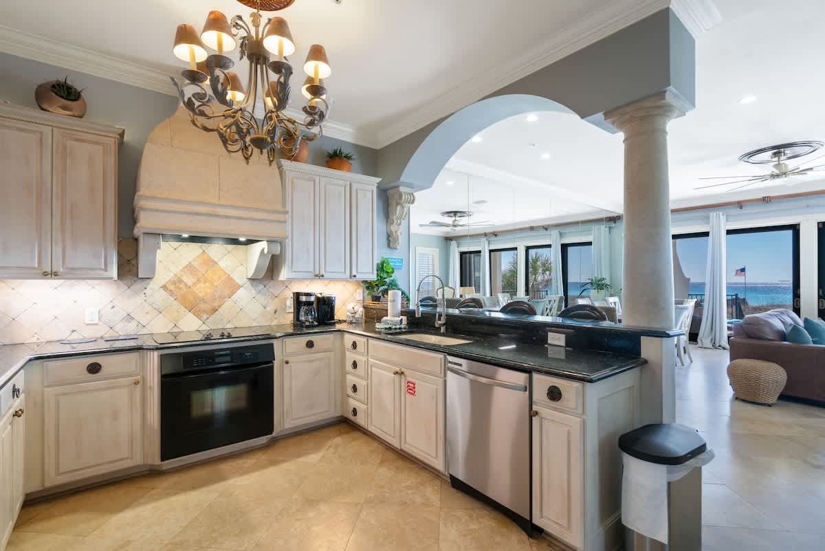 A spacious kitchen with light wood cabinets, dark granite countertops, and a decorative tile backsplash, opening up to a living area with ocean views through large windows. A large chandelier hangs centrally.