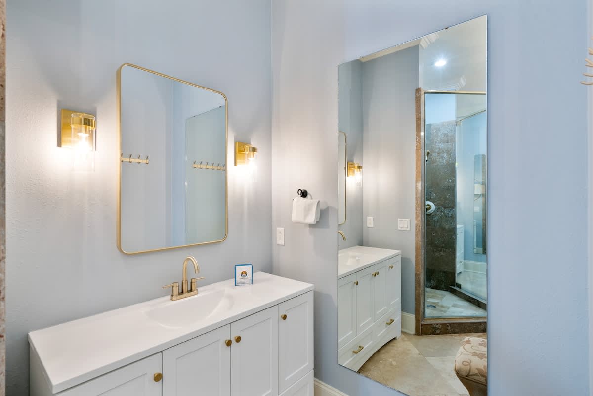 A bright bathroom vanity with a white cabinet, gold faucet, and a large gold-framed mirror, complemented by gold wall sconces and a full-length mirror reflecting the shower area.