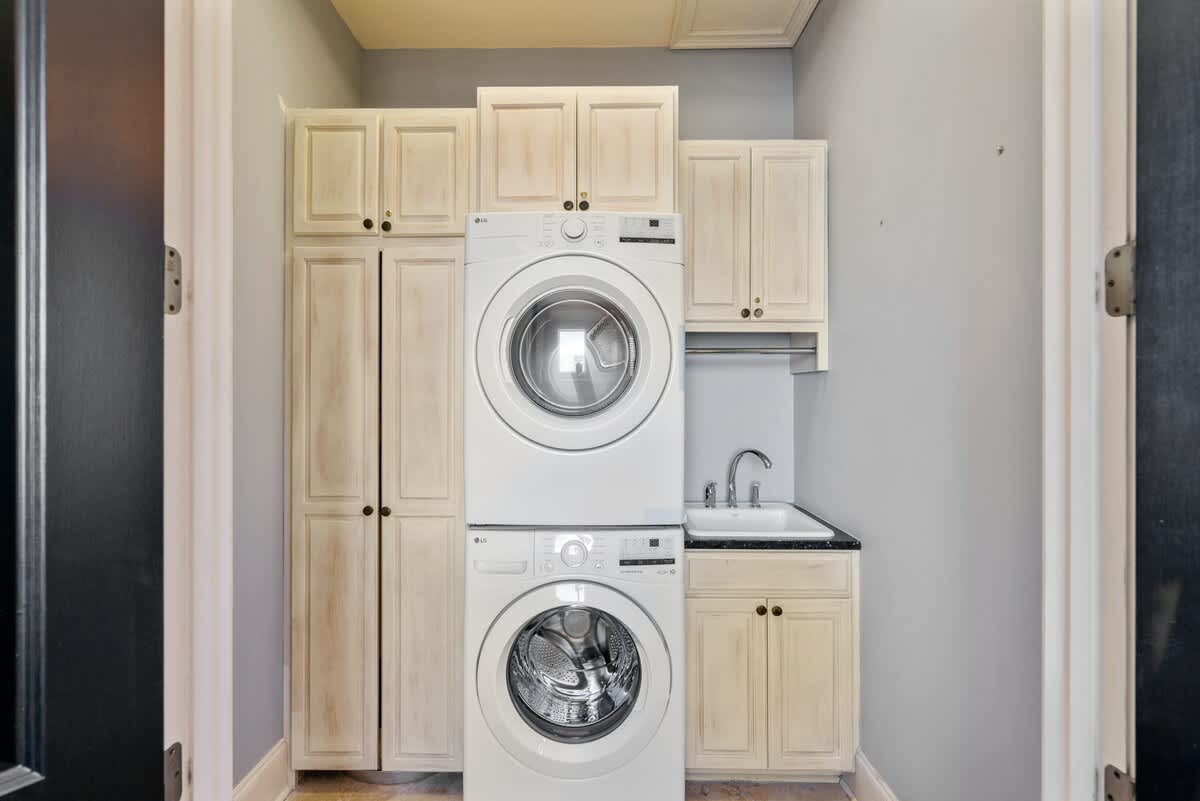 Compact laundry area with stacked white washer and dryer units, surrounded by light-colored wooden cabinetry. A small utility sink with a black countertop is located to the right.