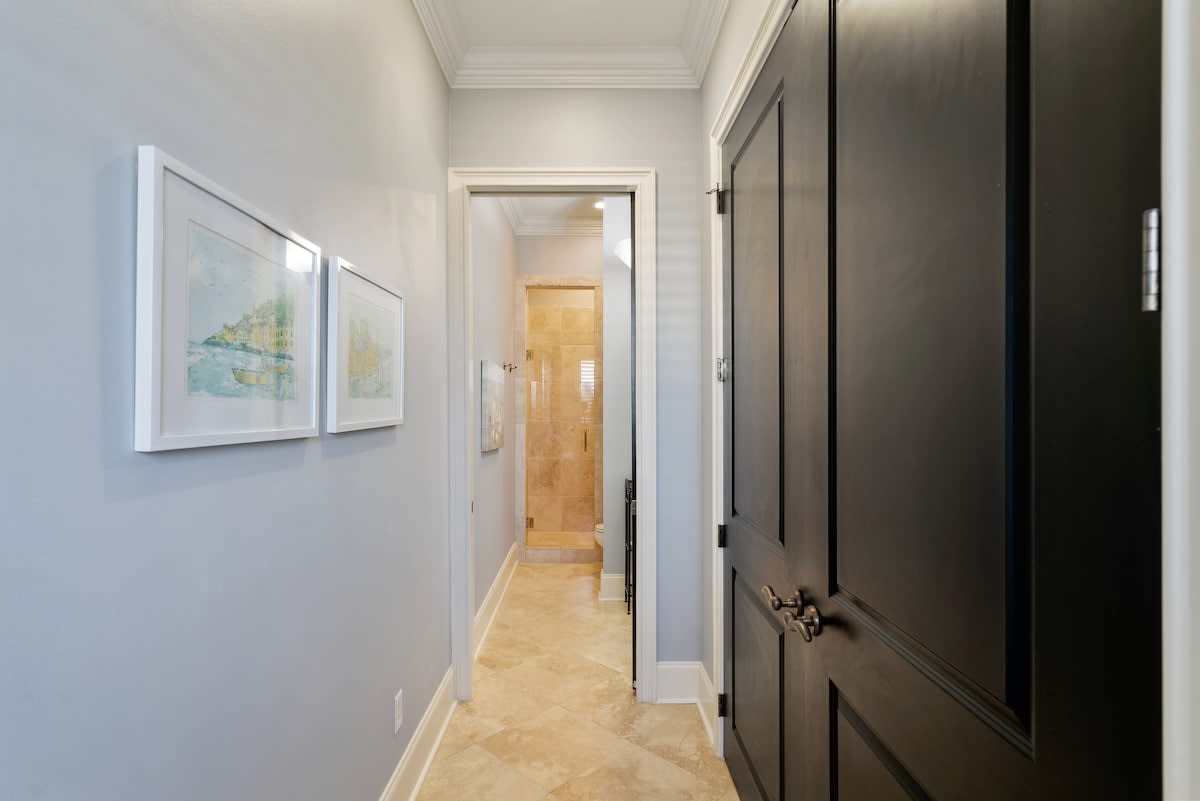 Interior hallway with light blue walls and travertine tile flooring, featuring framed artwork on the left wall. The hallway leads towards a bathroom with a glass-enclosed shower visible in the distance.