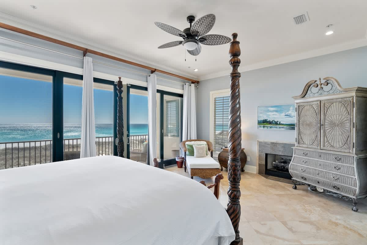 A master bedroom with light blue walls and travertine flooring, featuring a dark wood four-poster bed, a decorative armoire, and a fireplace, with glass doors offering panoramic ocean views.