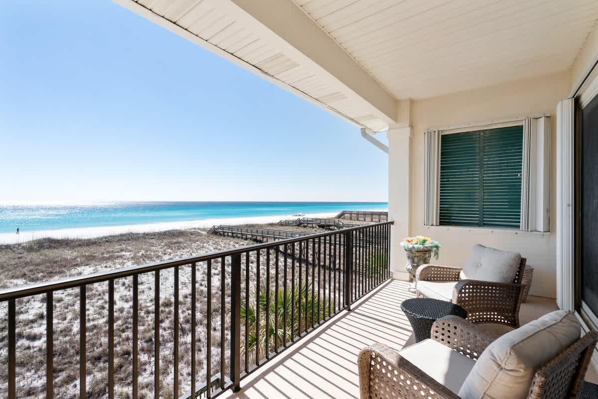 Covered balcony with black metal railing, featuring wicker chairs and a small table, overlooking the pristine white sand beach, turquoise ocean, and a wooden boardwalk. A window with louvered shutters is visible on the right.