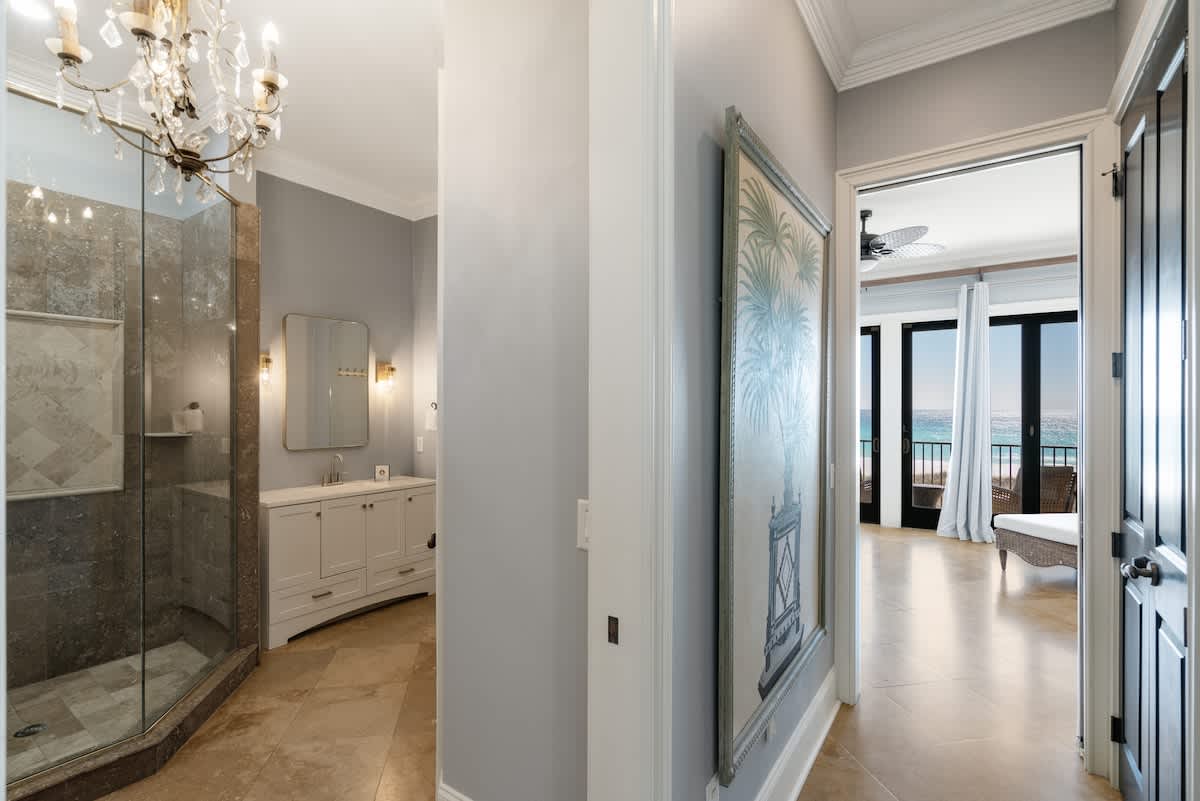 A hallway with light blue walls and travertine flooring, showcasing a large framed artwork and leading to a bathroom with a glass shower and vanity, and a bedroom with a balcony and ocean view.