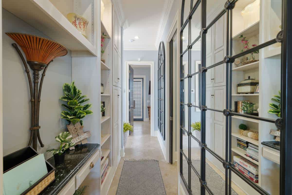 A narrow hallway with light-colored tile flooring, featuring built-in white shelving units adorned with decorative items and plants. A large arched mirror with a grid pattern reflects the opposite wall.