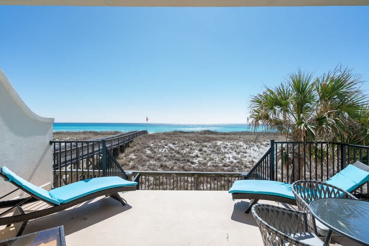 An outdoor balcony with two blue chaise lounge chairs and a small table, overlooking a wooden boardwalk, sand dunes, and the turquoise ocean under a clear blue sky.
