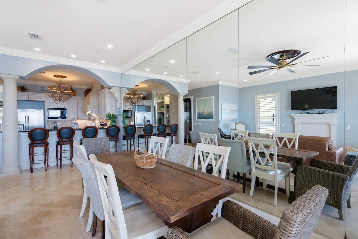 A large dining area with a rustic wooden table and various chairs, featuring a mirrored wall reflecting the living space and an open view into a well-appointed kitchen with bar seating.