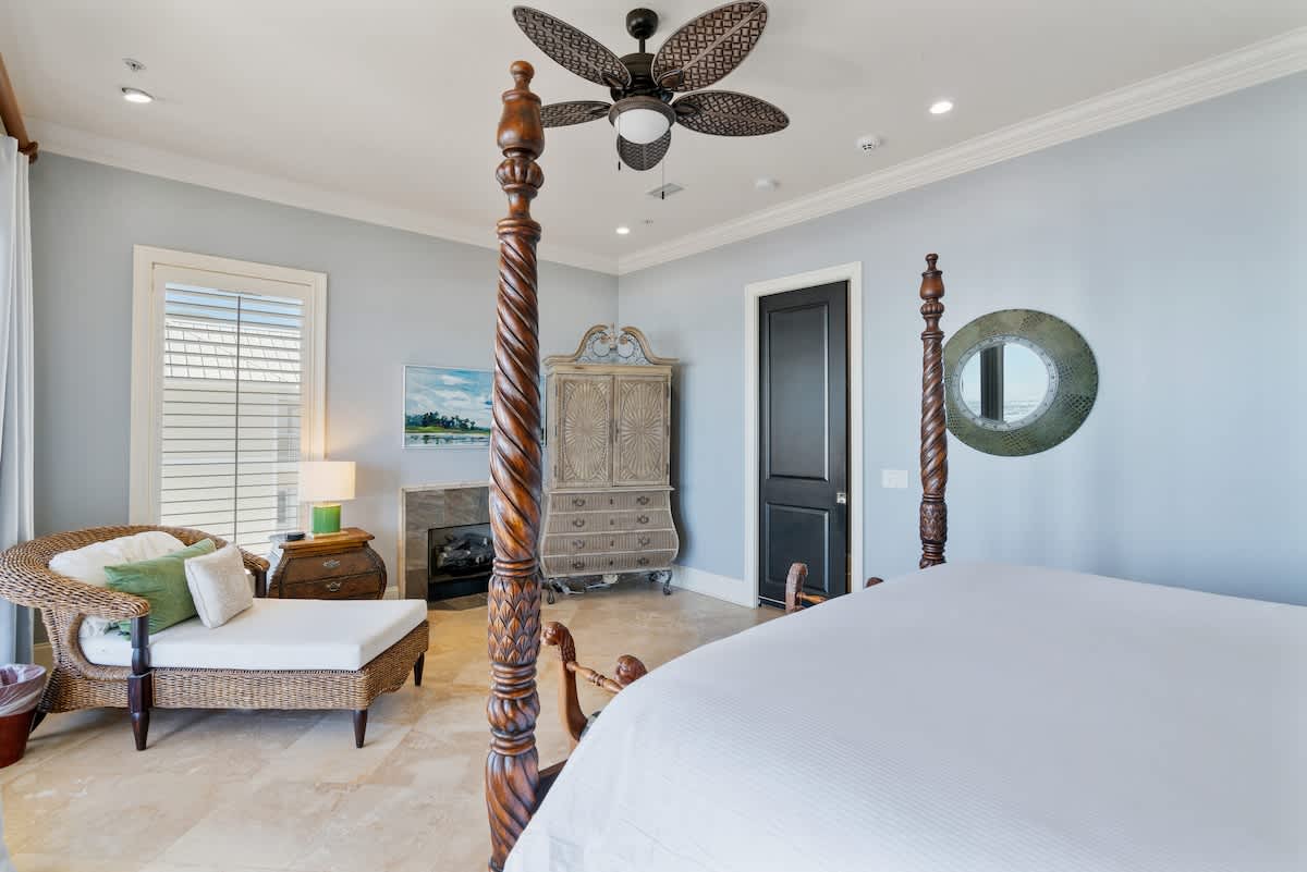 Spacious bedroom with light blue walls and travertine tile flooring, featuring a grand dark wood four-poster king bed with white bedding. A wicker chaise lounge and an antique-style armoire are also present, with a ceiling fan overhead.