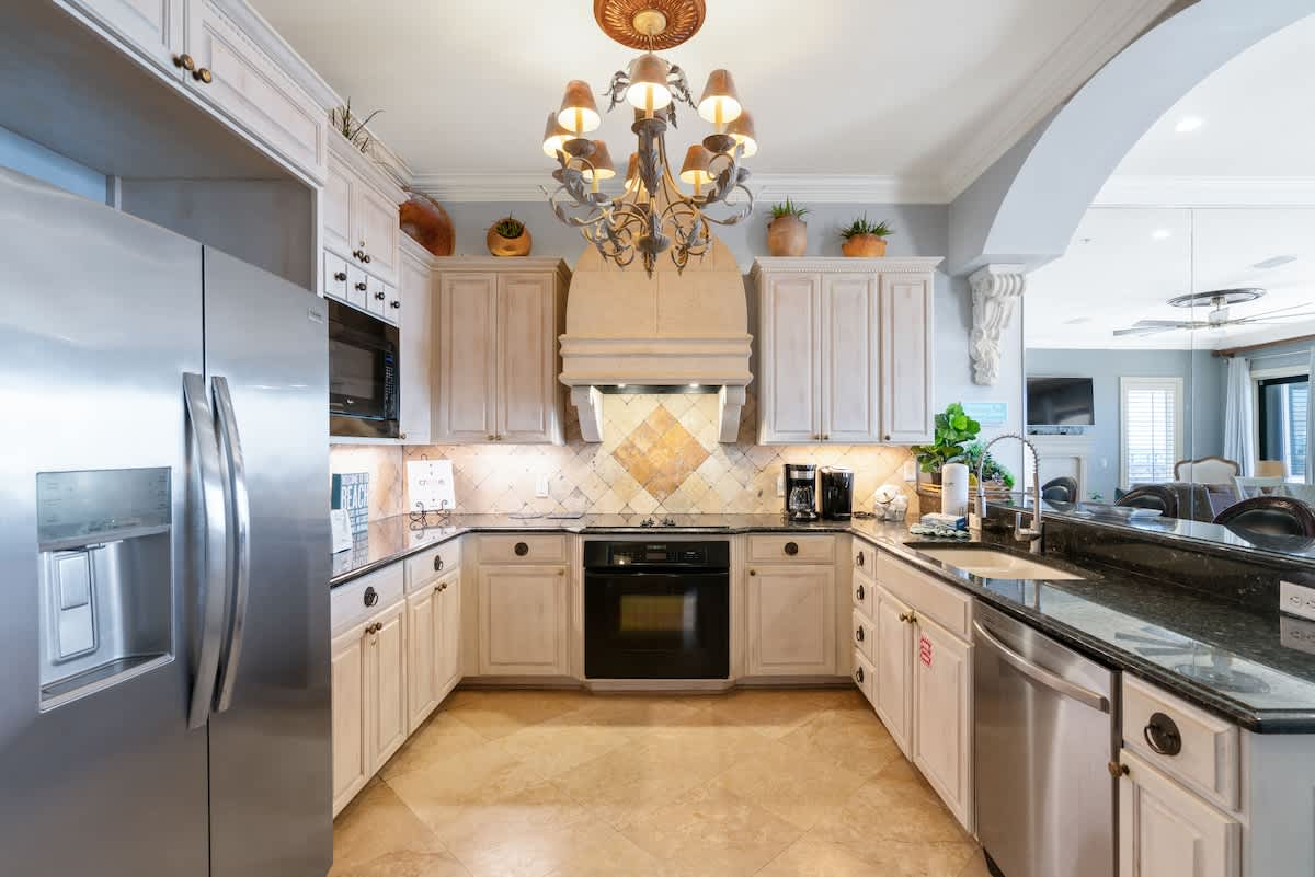 A well-lit kitchen featuring light-colored cabinetry, dark granite countertops, and a diamond-patterned tile backsplash. A large stainless steel refrigerator is visible on the left, and a decorative chandelier hangs above.