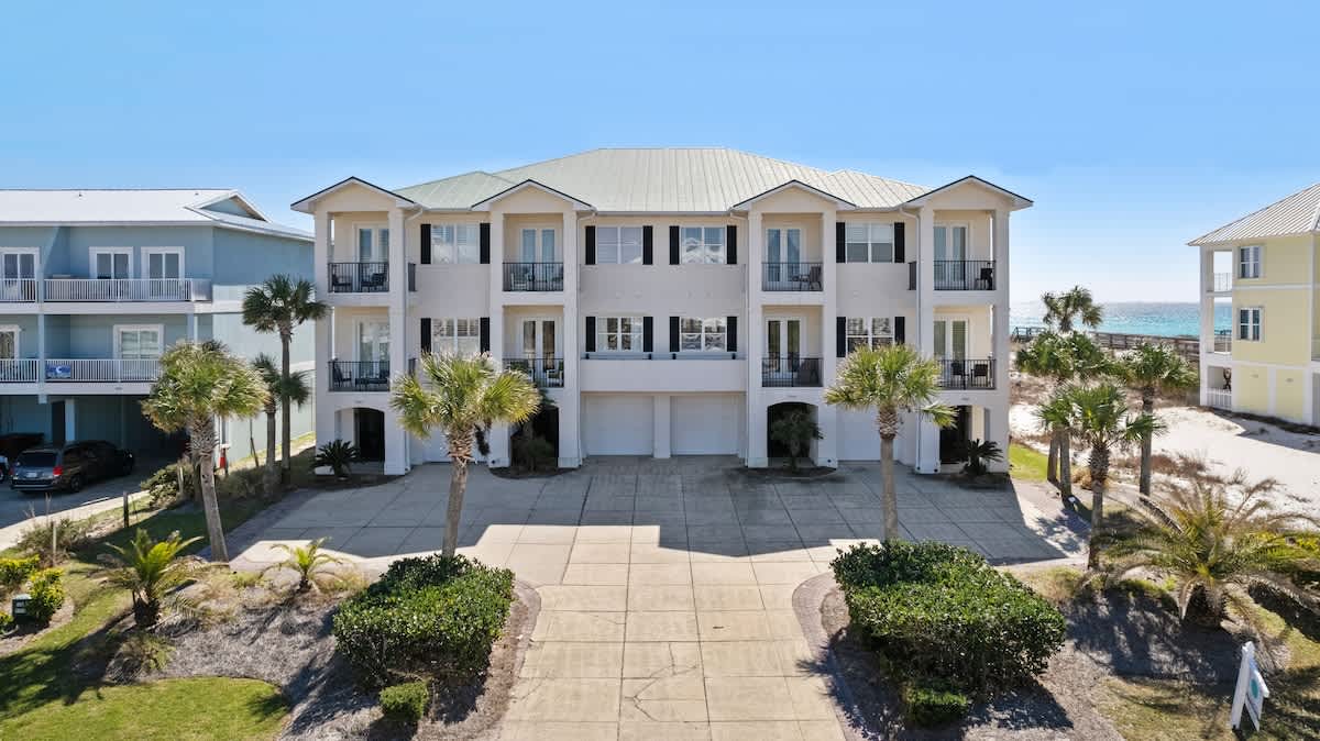 The front exterior of a large, multi-story townhouse building with a white facade, green roof, multiple balconies, and a paved driveway surrounded by palm trees and landscaping.