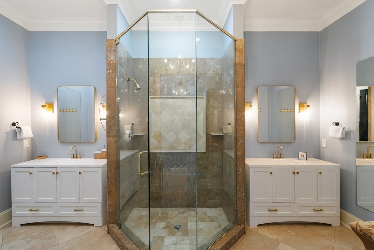 A spacious bathroom with light blue walls and travertine flooring, featuring a central glass-enclosed corner shower with tiled walls, flanked by two white vanities with gold-framed mirrors and sconces.
