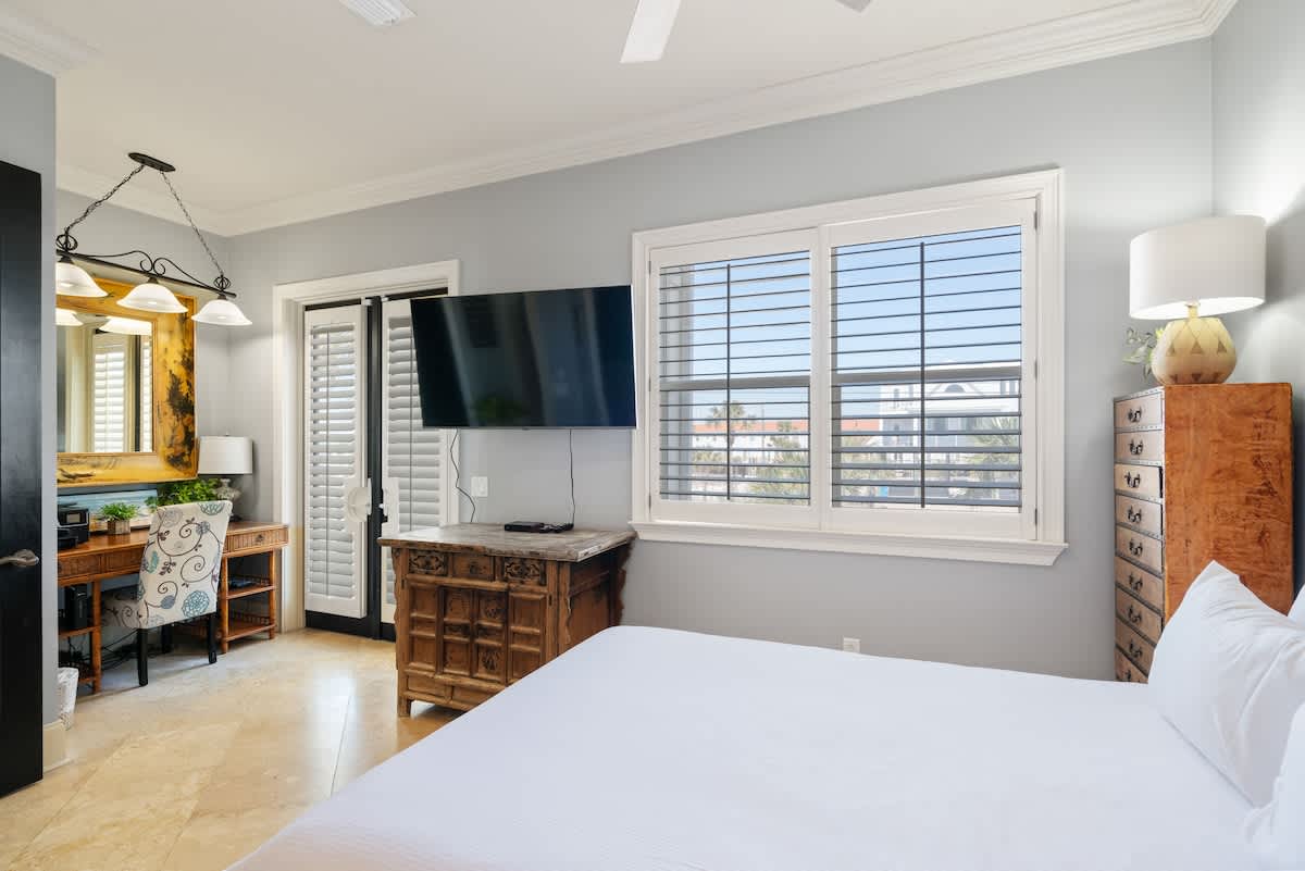 A bright bedroom with light blue walls and tile flooring, featuring a queen bed, a wall-mounted TV, and a dedicated desk area with a patterned chair. A large window with white plantation shutters offers natural light.