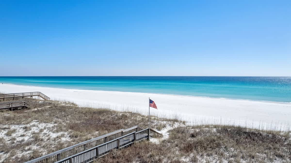 A pristine white sand beach stretches along the vibrant turquoise ocean under a bright blue sky, with a wooden boardwalk and an American flag visible in the distance.