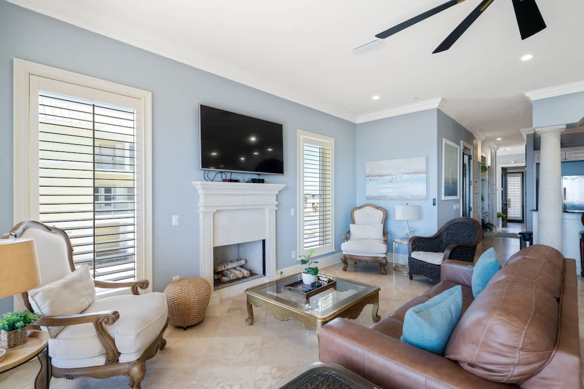 A spacious living room with light blue walls, tiled floors, a brown leather sofa, and various armchairs, centered around a gold coffee table and a stone fireplace, with natural light from windows.