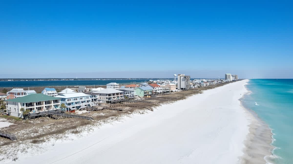 An aerial view of a long stretch of white sand beach and turquoise ocean, with a line of beachfront houses and buildings nestled behind sand dunes.