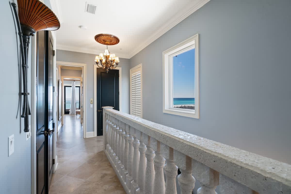 A bright upper-level hallway with light blue walls, travertine tile flooring, and a white balustrade, featuring a window with a stunning ocean view and a decorative chandelier.