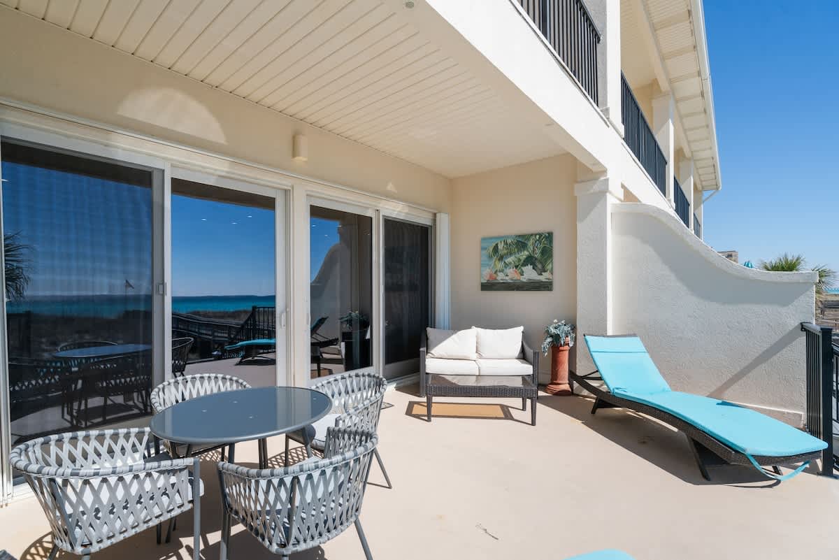 A spacious outdoor patio with a gray round dining table and chairs, a small sofa, and a blue chaise lounge, offering a partial view of the ocean through large sliding glass doors.