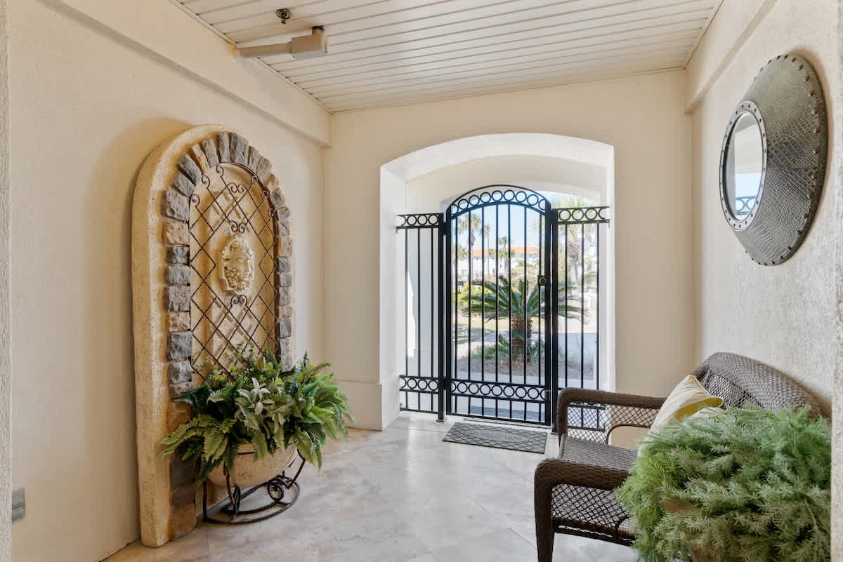 Elegant foyer with light tile flooring, featuring a decorative arched stone and metal wall accent with lush potted plants. A wicker bench provides seating, and a black wrought iron gate leads to an outdoor area with visible palm trees.